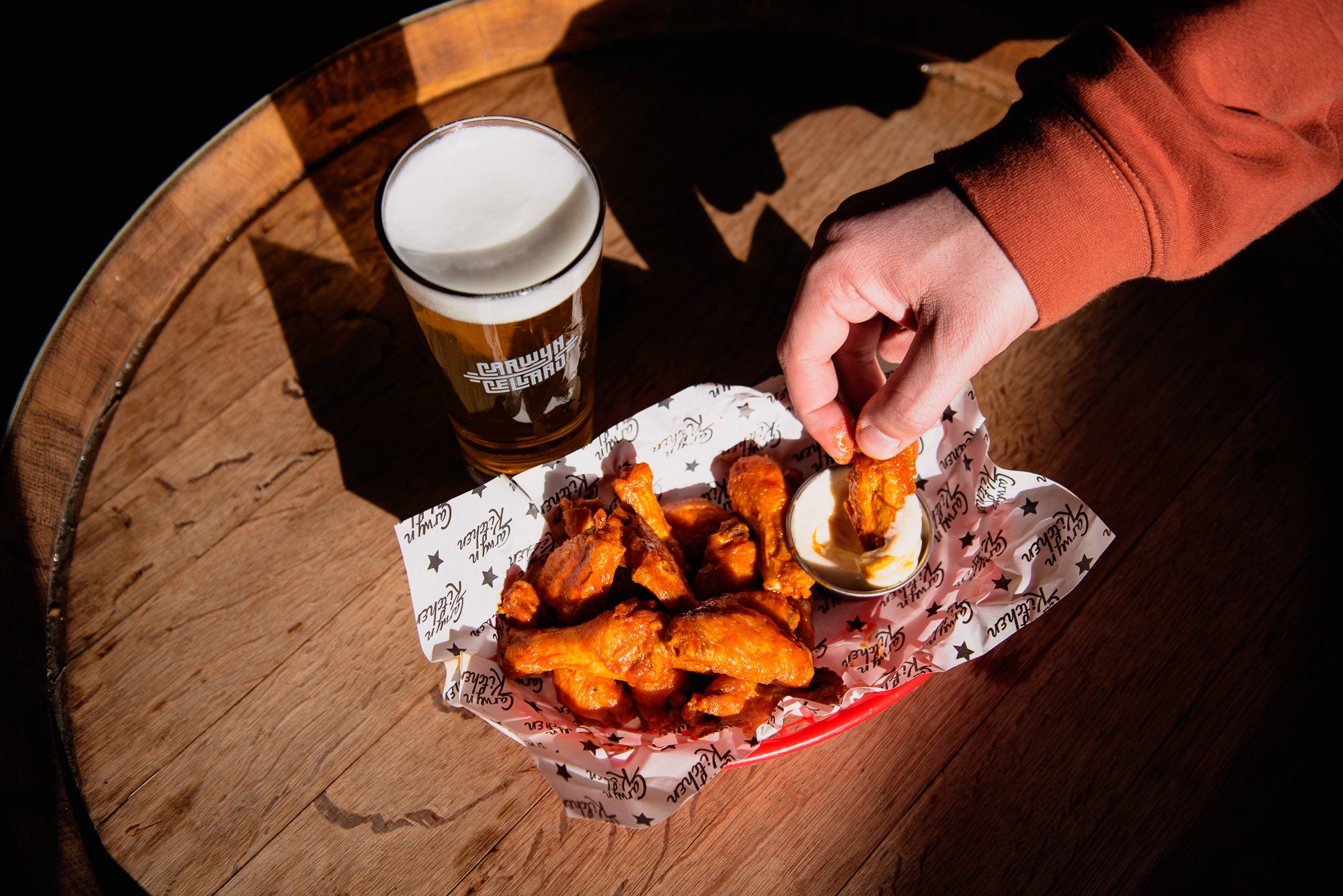 A person's hand dipping a piece of fried chicken into a small sauce cup, with a basket of wings on branded paper and a pint of beer on a wooden table.